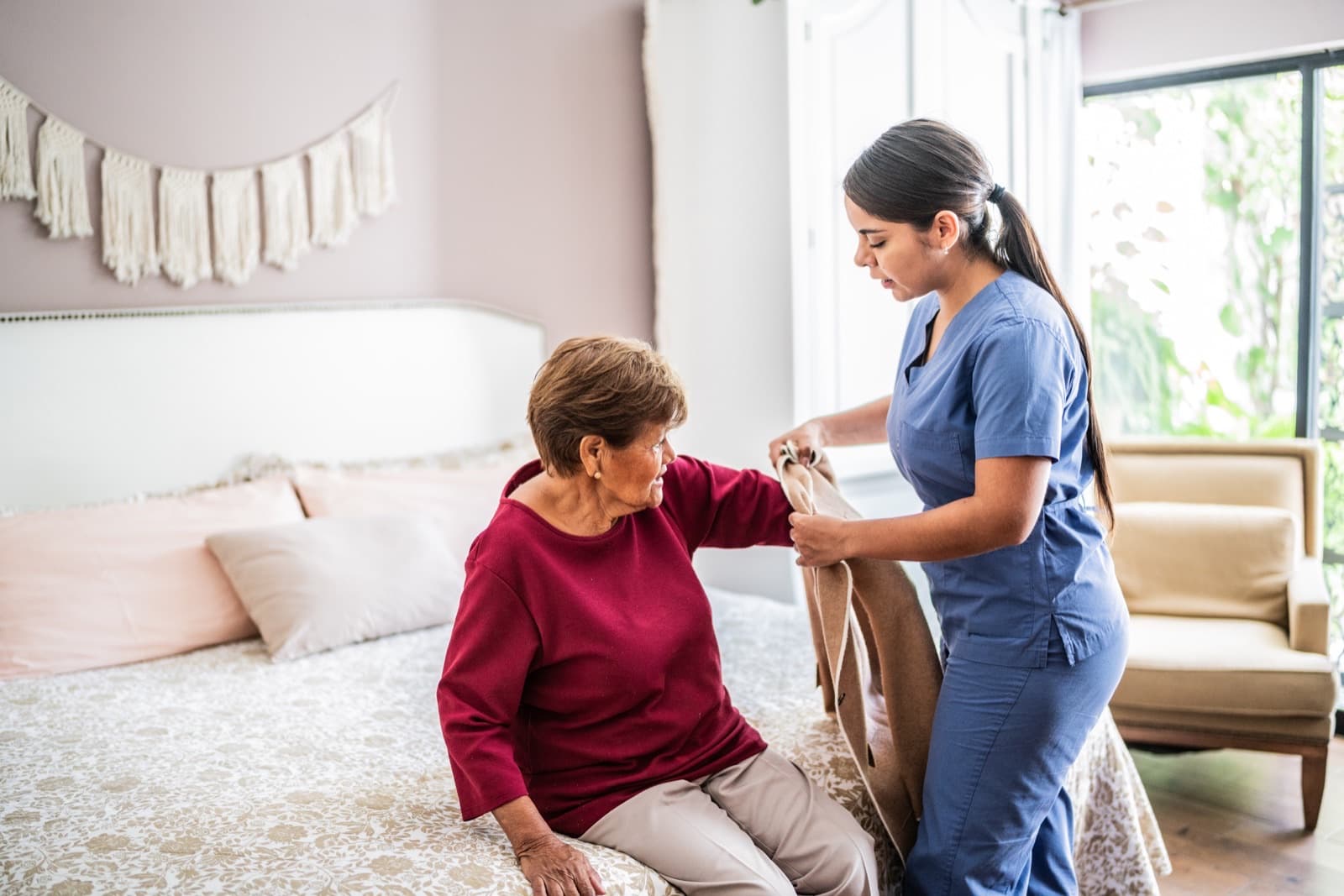 Supportive companion helping an older adult with getting dressed in a bright home bedroom