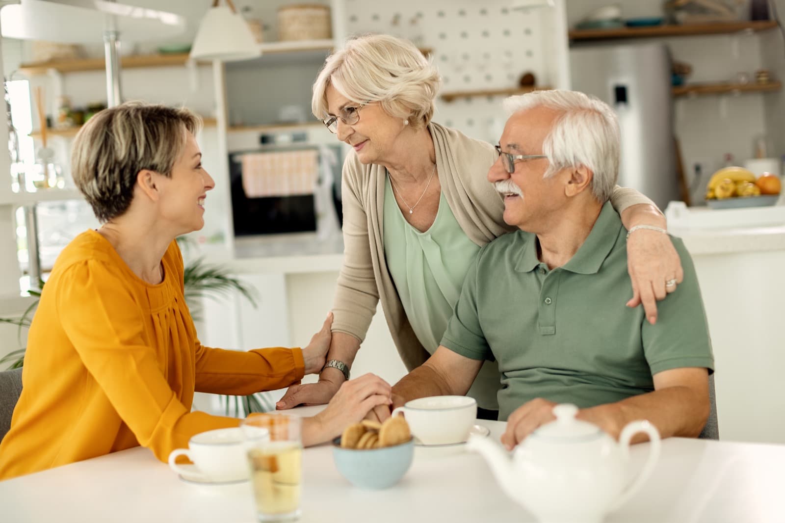 Caregiver sharing tea with an older couple at a kitchen table in a warm home
