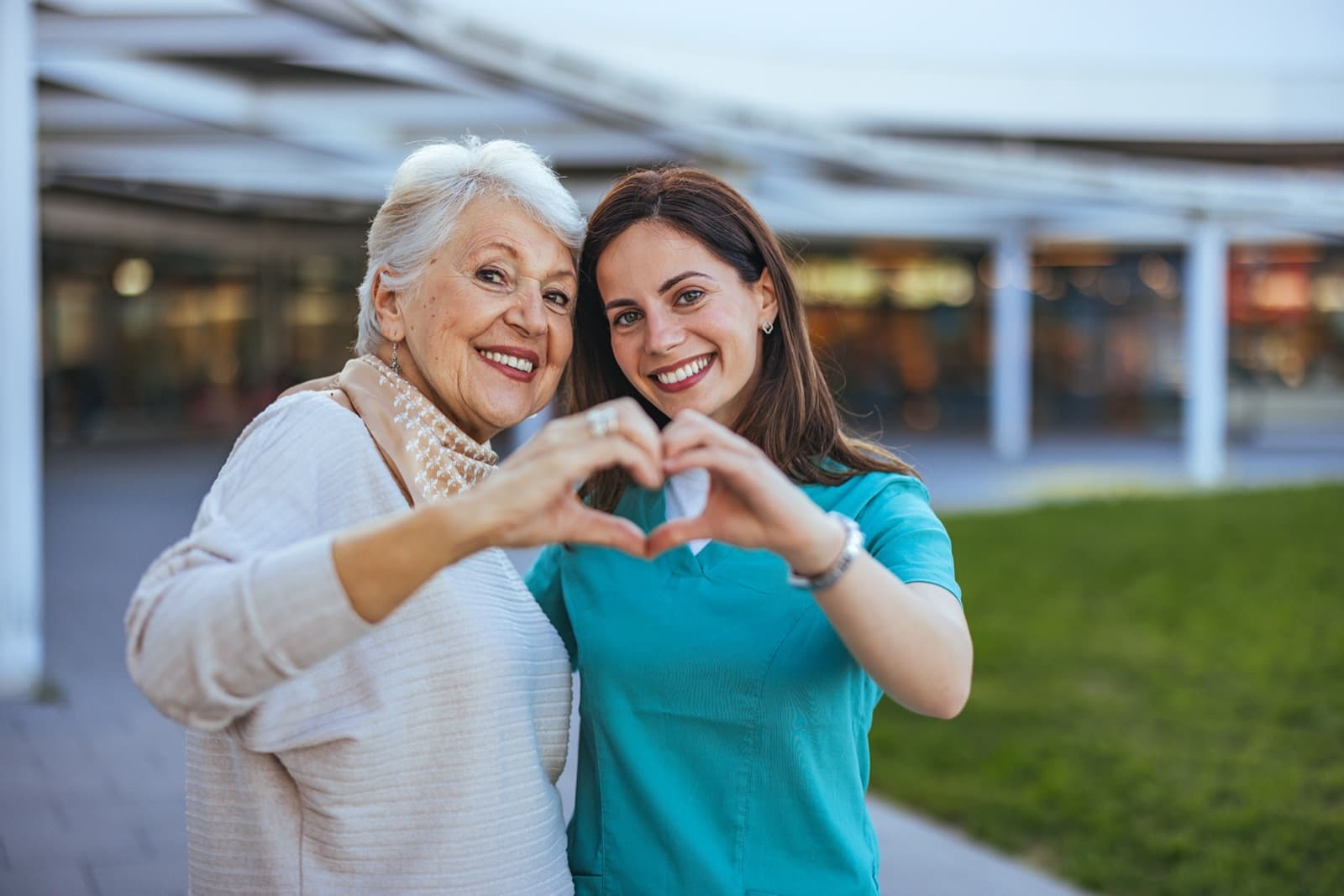 Older woman and younger companion smiling together outdoors, both making a heart shape with their hands