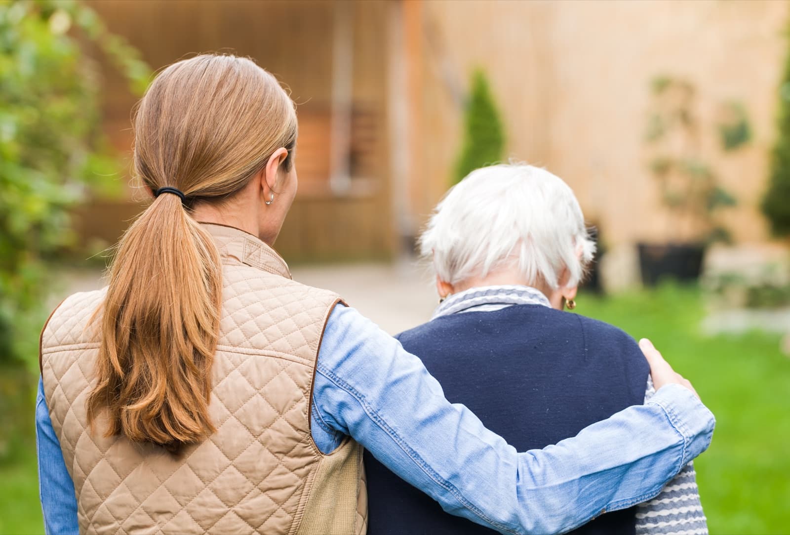 Younger woman walking outdoors with her arm around an older woman's shoulder on a sunny path