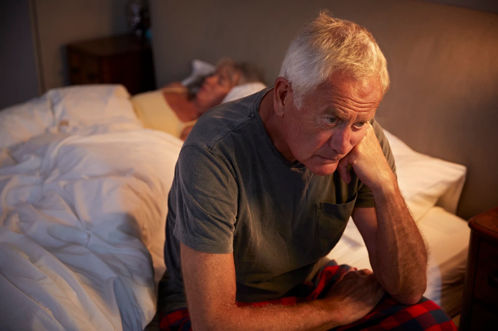 Older adult sitting on the edge of a bed at night while a partner sleeps nearby